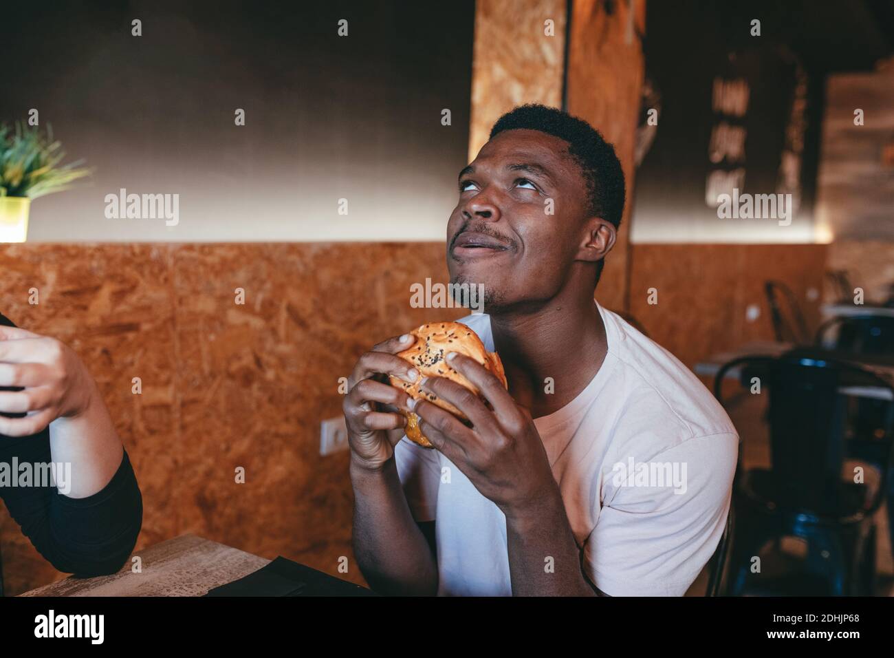 Bon mâle afro-américain qui mange un délicieux hamburger et qui apprécie le goût tout en regardant vers le haut et en faisant rouler les yeux Banque D'Images
