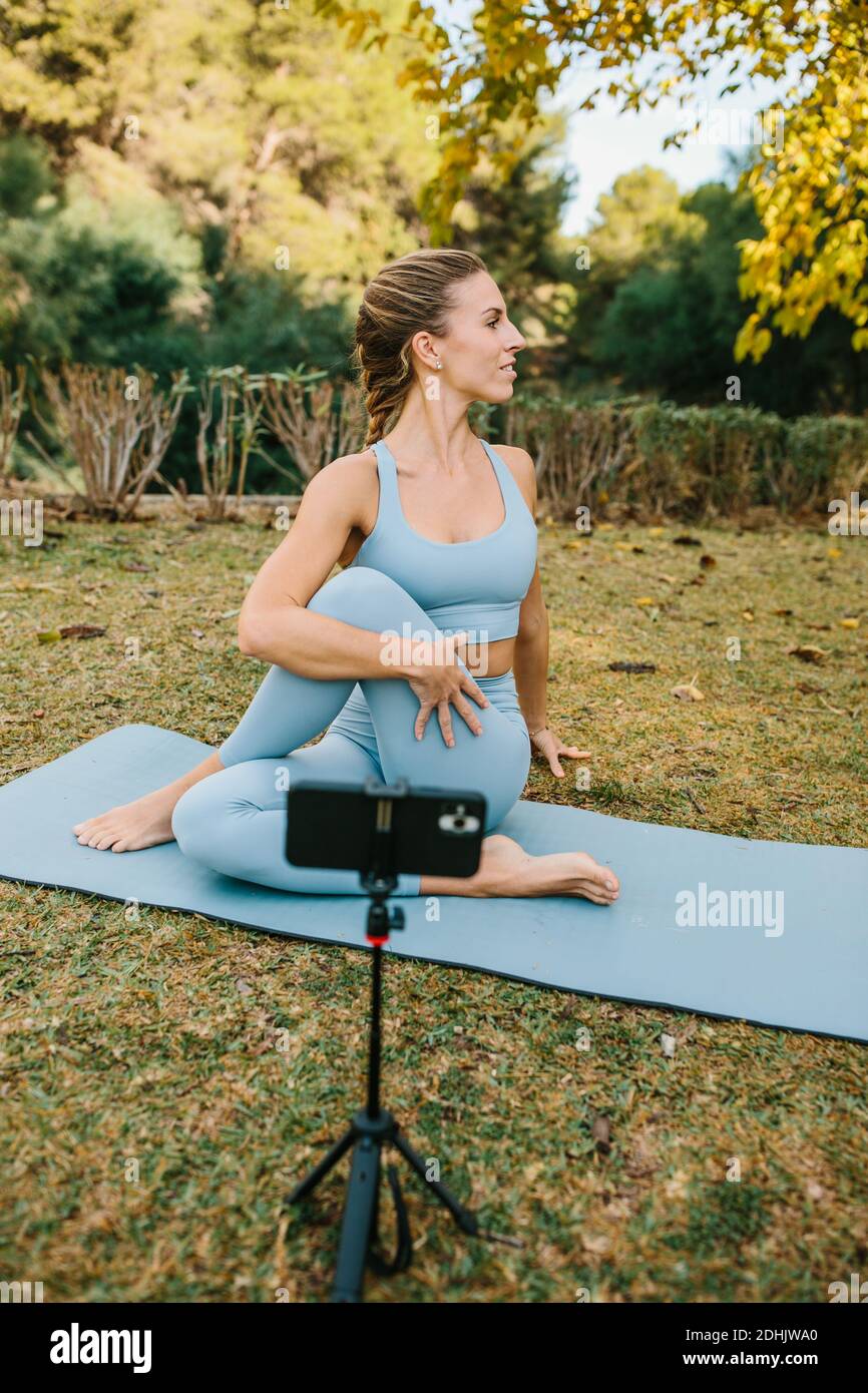 Flexible femme pratiquant le yoga et l'enregistrement vidéo sur smartphone pour Blog tout en étant assis à Ardha Matsyendrasana sur tapis dans le parc Banque D'Images