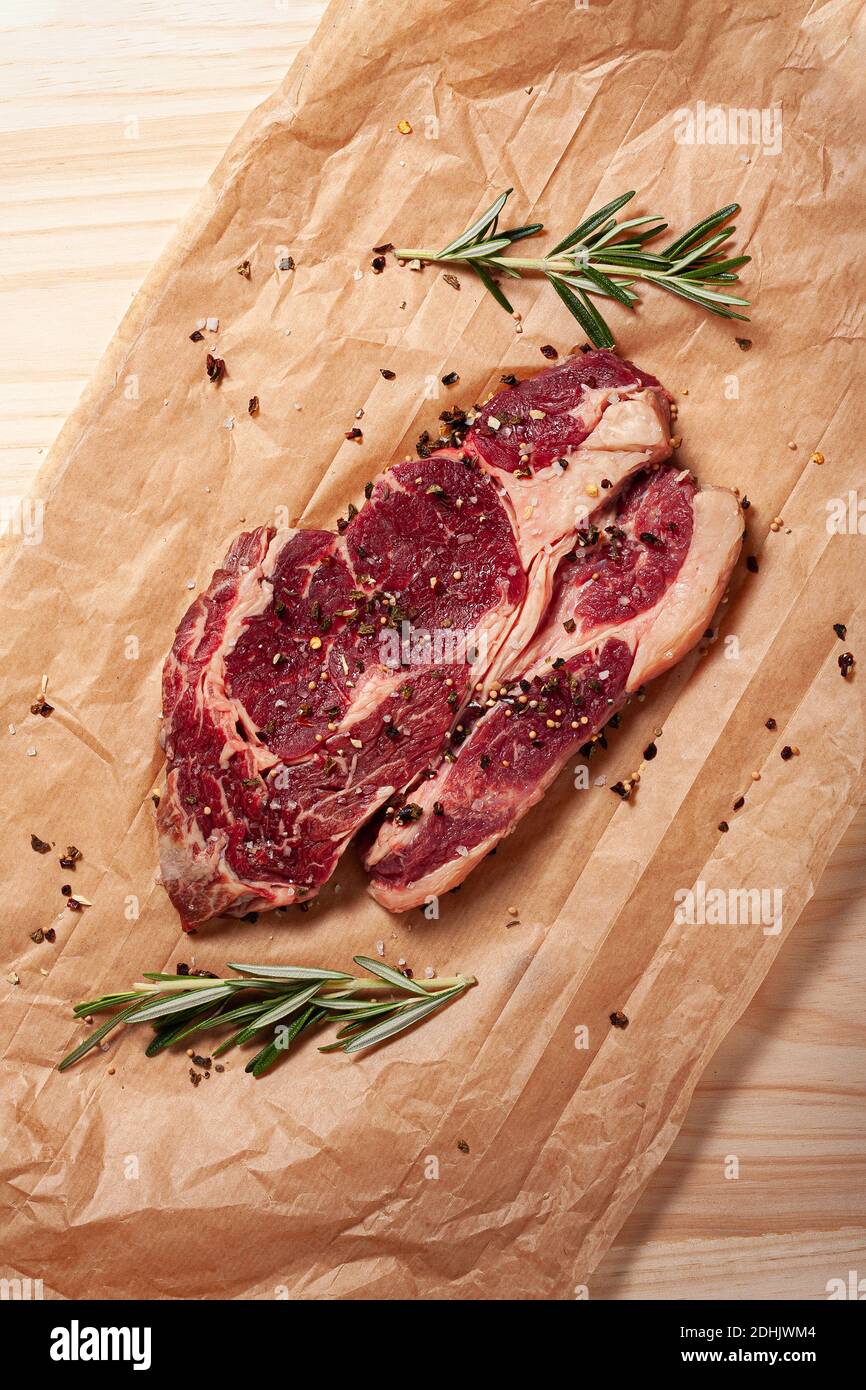 Vue de dessus du steak de bœuf aux yeux de côtes juteuses fraîchement non cuites avec du romarin frais et du poivre sur une feuille de papier de cuisson sur la table Banque D'Images