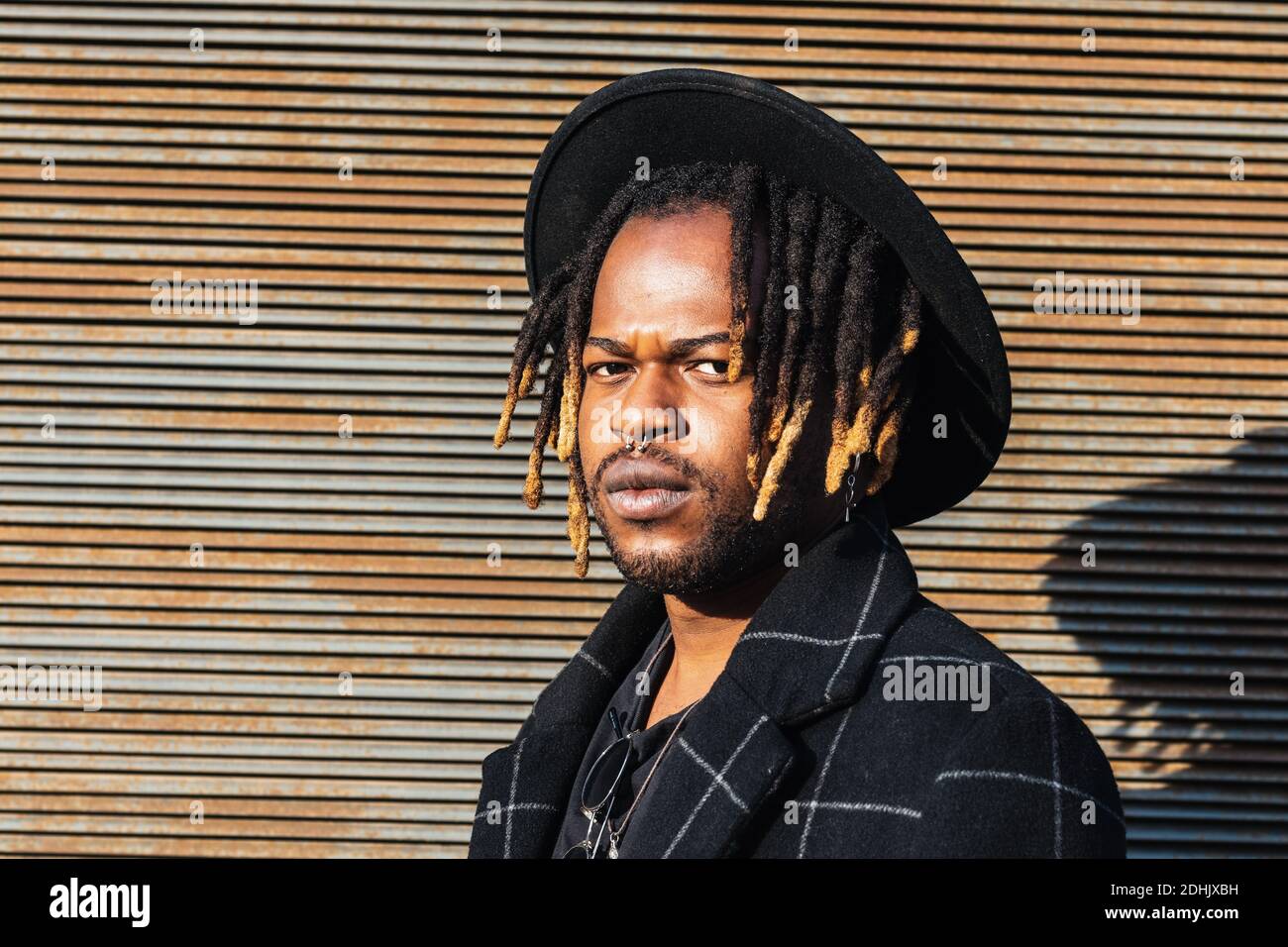 Homme noir moderne pour adultes avec des dreadlocks portant un chapeau et élégant manteau regardant l'appareil photo contre le mur de rue en plein soleil Banque D'Images