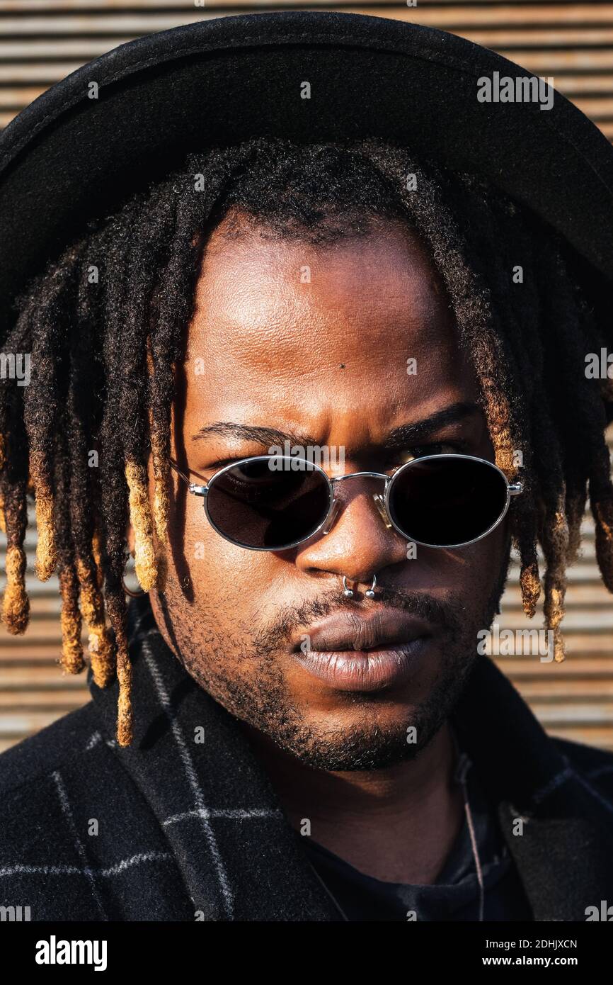 Homme noir moderne pour adultes avec des dreadlocks portant un chapeau, des lunettes de soleil et un manteau élégant regardant l'appareil photo contre le mur de rue en plein soleil Banque D'Images