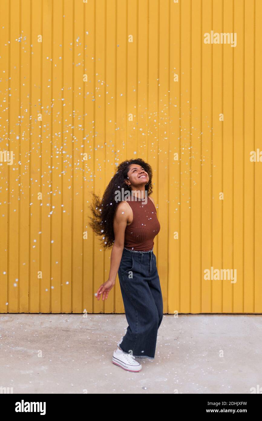 Joyeuse afro-américaine avec des confettis blancs de diffusion de cheveux bouclés en ville tout en s'amusant et en regardant Banque D'Images