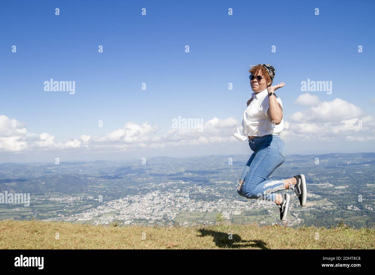 une belle jeune femme latino-américaine en plein air avec des lunettes de soleil heureux et de sauter dans une journée d'été avec vue de la ville dominicaine distante Banque D'Images