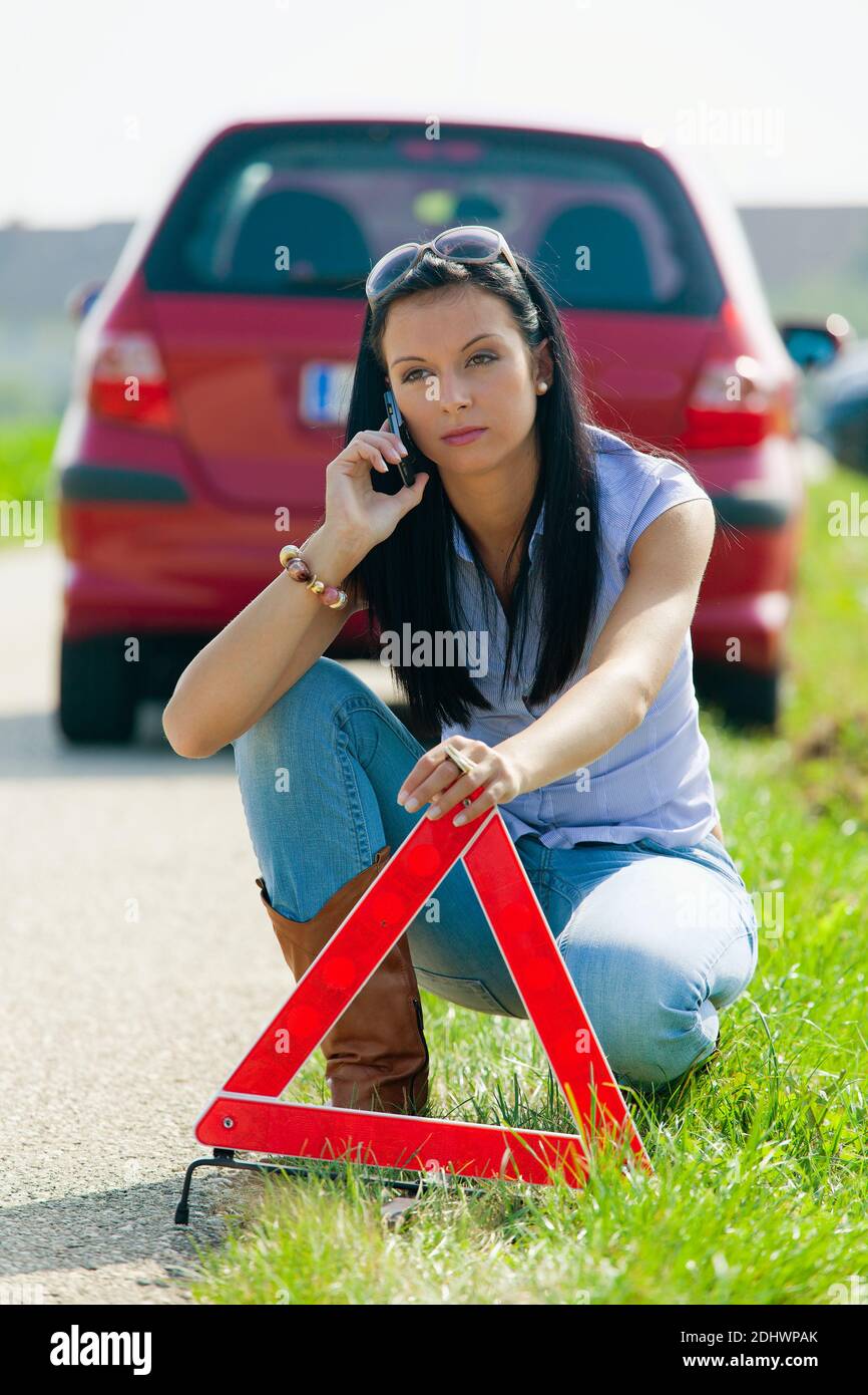 Junge Frau mit einer Reifenpanne am Auto, Warnkreuz wird aufgestellz, Banque D'Images