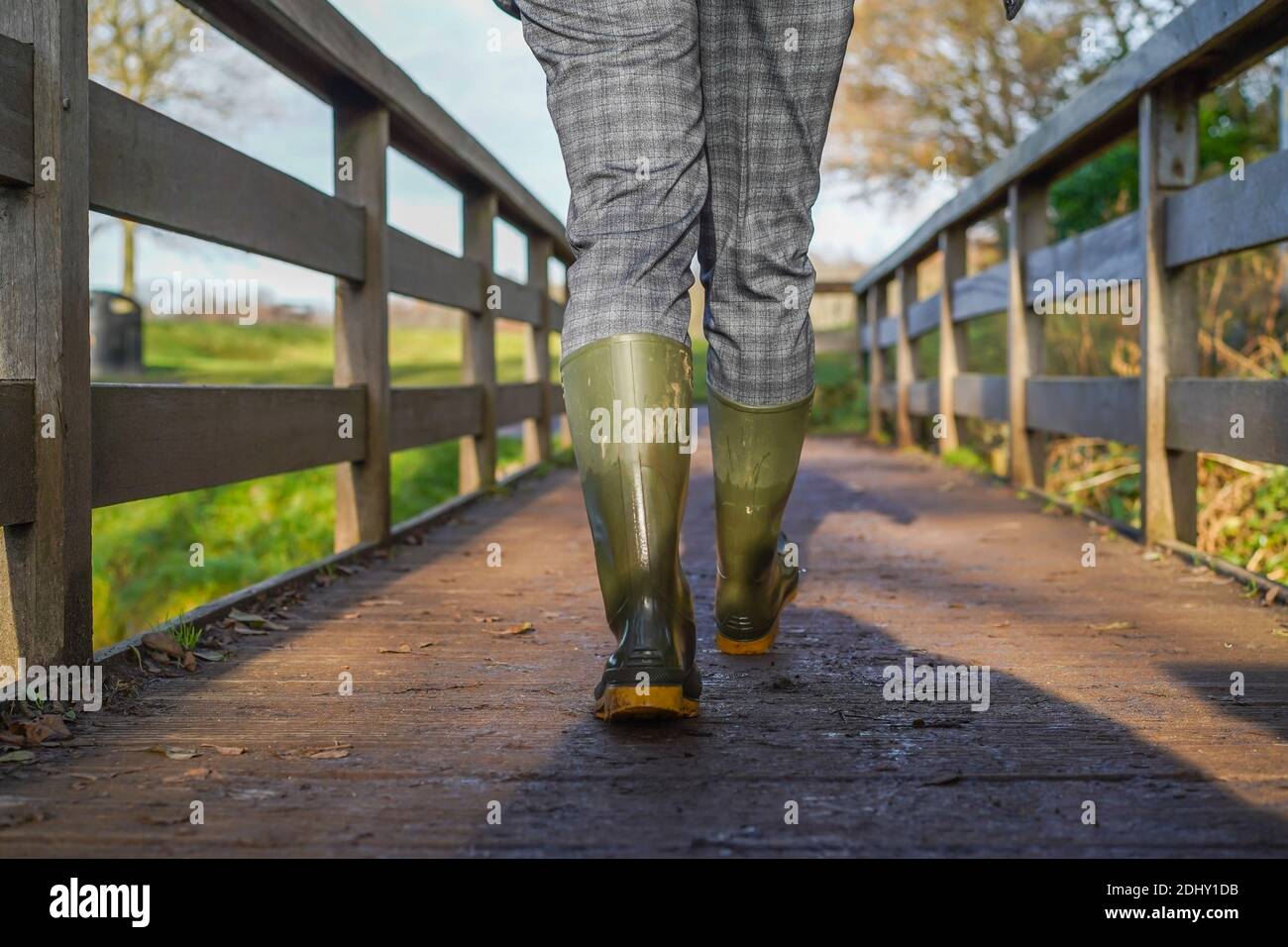 Vue arrière à angle bas de la femme britannique en bottes/bottes wellington, isolée en plein air marchant sous le soleil hivernal traversant une passerelle en bois. Banque D'Images