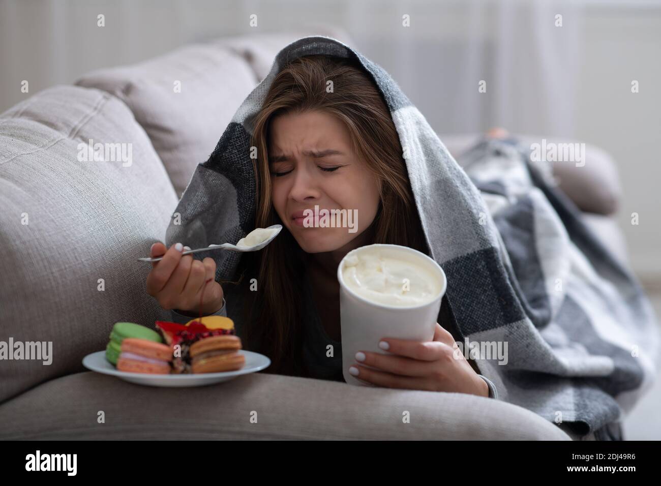 Bouleverser la jeune femme qui fait face à la dépression en mangeant de la crème glacée et des bonbons sous la couverture sur le canapé Banque D'Images