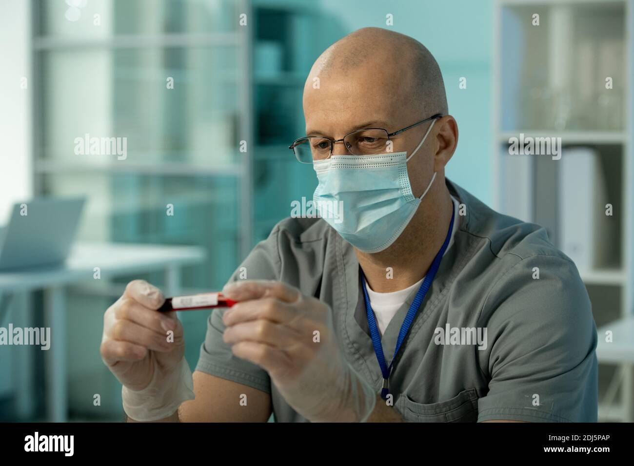 Homme bald mature scientifique dans le masque de protection et les gants regardant à l'échantillon de sang dans une petite fiole pendant la recherche scientifique dans laboratoire chimique Banque D'Images