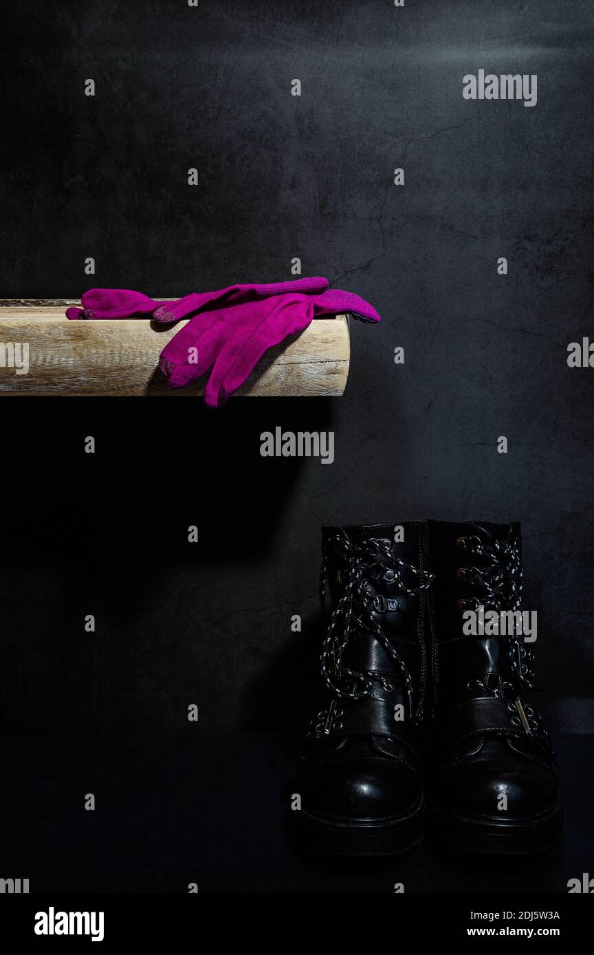 Image d'une paire de gants roses sur une étagère en bois sur fond sombre. Sous la tablette se trouve une paire de bottes de motard noires sur un sol noir Banque D'Images