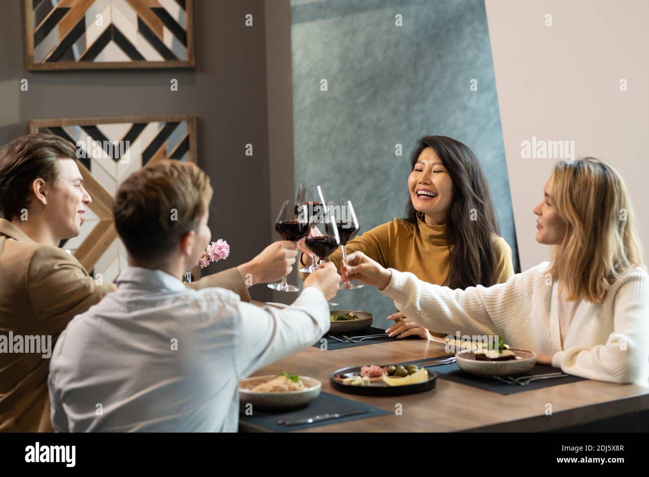 Deux jeunes couples interculturels en vêtements décontractés élégants se clinking avec des lunettes de vin rouge servi à table par dîner dans le luxe restaurant Banque D'Images