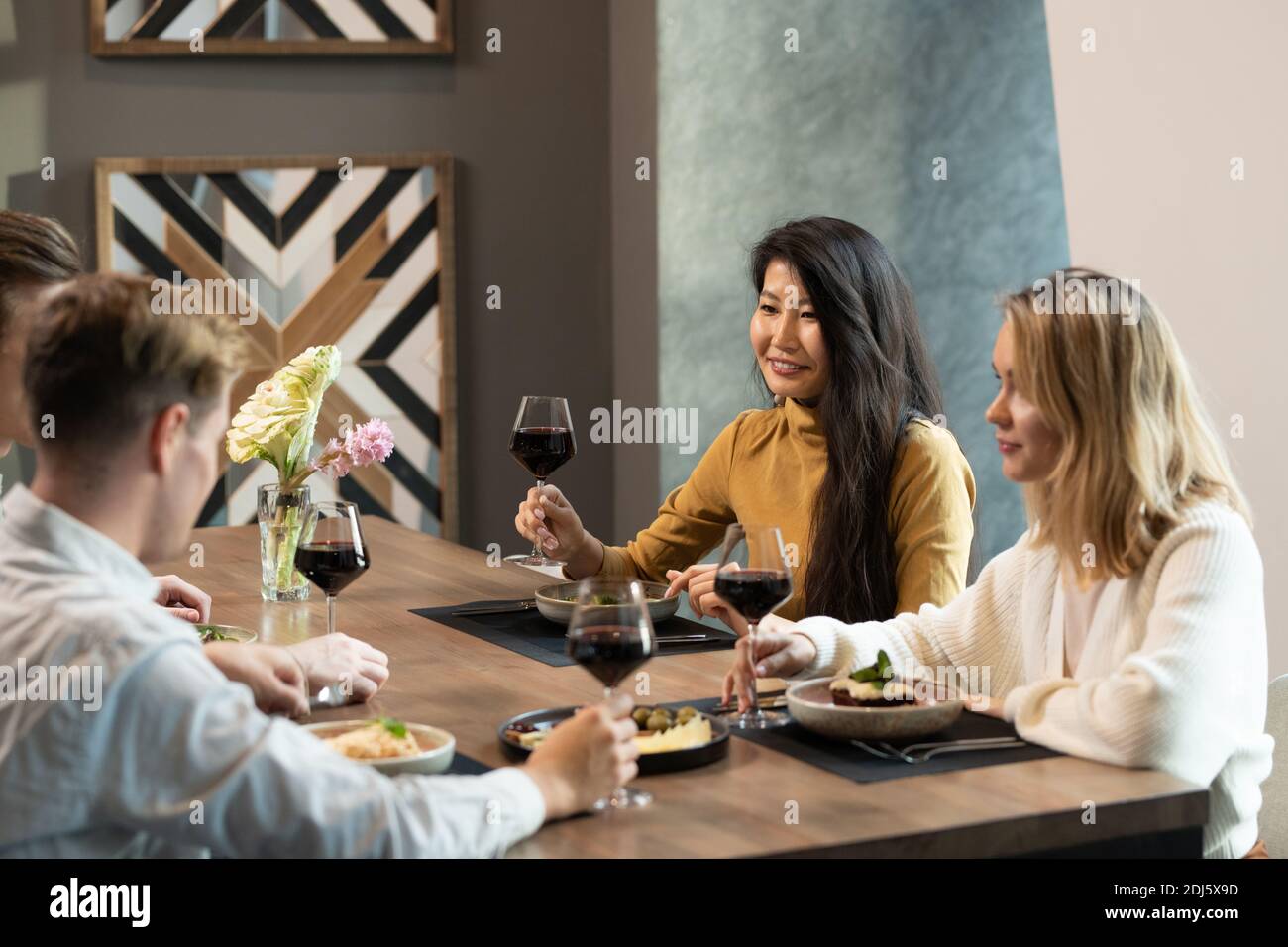 Jeune femme asiatique élégante avec un verre de vin rouge toast au dîner assis près d'une table servie dans le restaurant parmi ses amis Banque D'Images