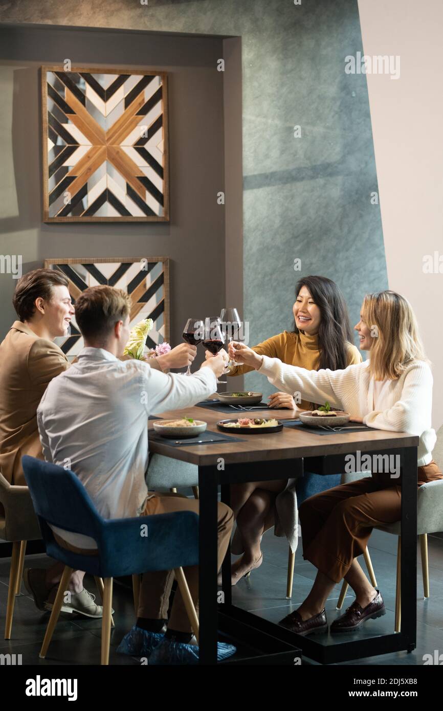 Deux jeunes couples gaies et amicaux en tenues décontractées et élégantes verres de vin rouge servis à table le soir restaurant Banque D'Images