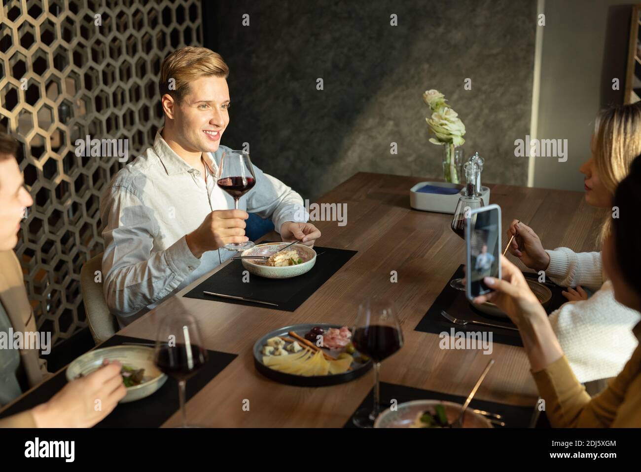 Joyeux jeune homme élégant avec un verre de vin rouge toast au dîner assis près d'une table servie dans un cadre moderne restaurant entre amis Banque D'Images