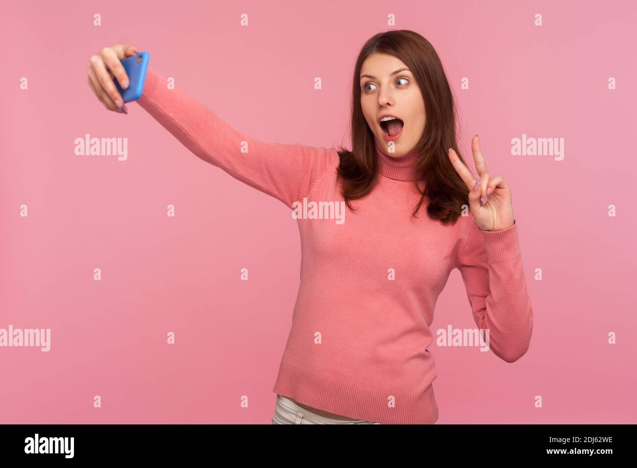 Blogueur féminine drôle avec des cheveux bruns montrant le geste de paix et le visage dupe en enregistrant la vidéo ou en faisant des photos sur smartphone. Prise de vue en studio isolée Banque D'Images