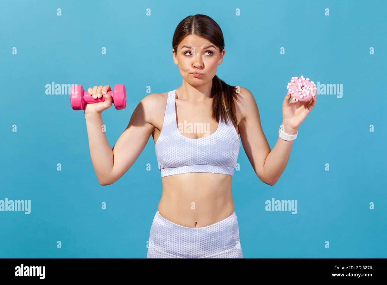 Femme athlétique confuse en blanc sportswear tenant dans les mains rose haltère et donut avec le glaçage rose, faisant le choix difficile, succomber à la séduction. Intérieur Banque D'Images