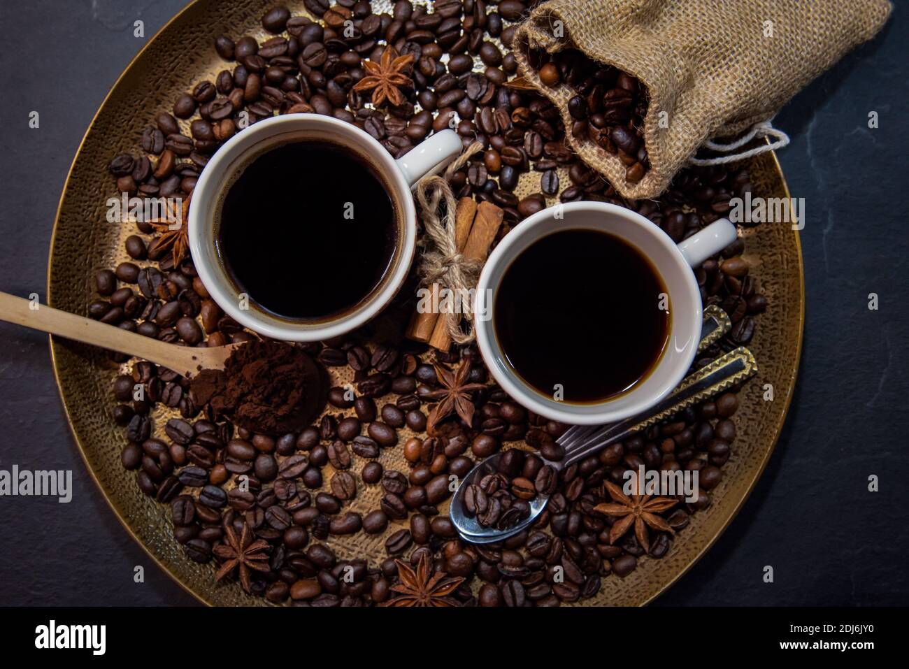 Vue de haut en bas de deux tasses de café sur un plateau de service doré entouré de café torréfié, café moulu, toile de jute avec des haricots rôtis et des épices. Banque D'Images