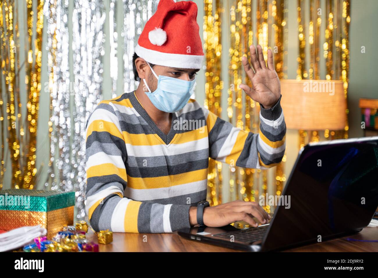Jeune homme dans un masque médical sur appel vidéo dans un ordinateur portable Pendant Noël ou la fête du nouvel an, avec un décor de fond et cadeaux en avant - concept Banque D'Images