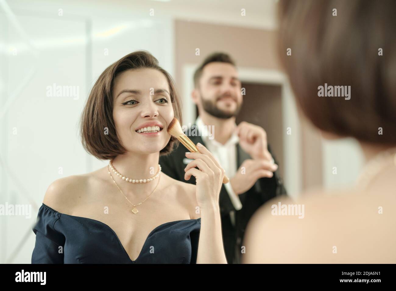 Bonne magnifique jeune femme appliquant le maquillage avec la brosse à l'avant de miroir tandis que son petit ami règle la manche de chemise Banque D'Images