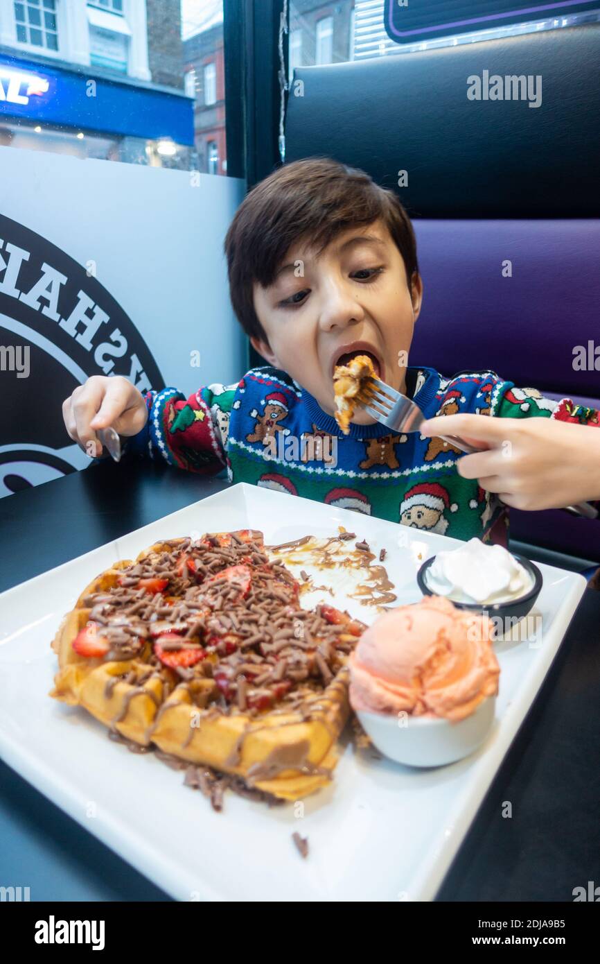 Un jeune garçon portant un pull de Noël mange une grande gaufre avec de la glace, des fraises et du chocolat. Banque D'Images