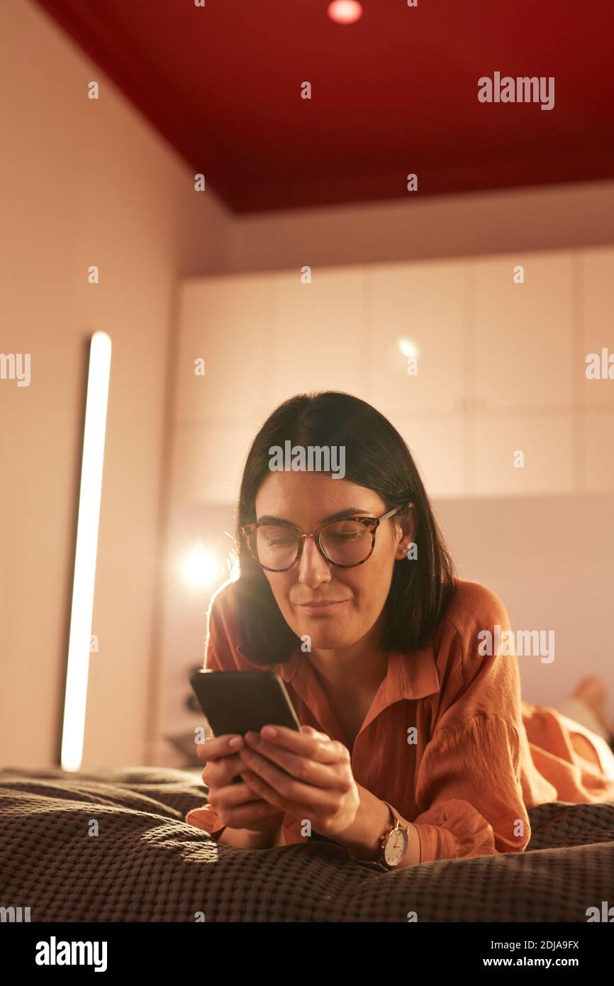 Portrait vertical d'une femme adulte contemporaine utilisant un smartphone pendant qu'elle est allongée sur le lit dans un intérieur minimal Banque D'Images