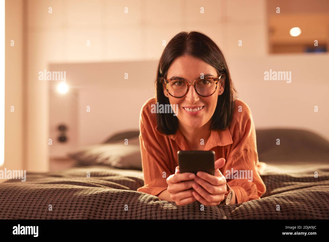 Portrait d'une femme adulte contemporaine utilisant un smartphone tout en étant couché sur le lit dans un intérieur minimal et souriant à l'appareil photo, espace de copie Banque D'Images