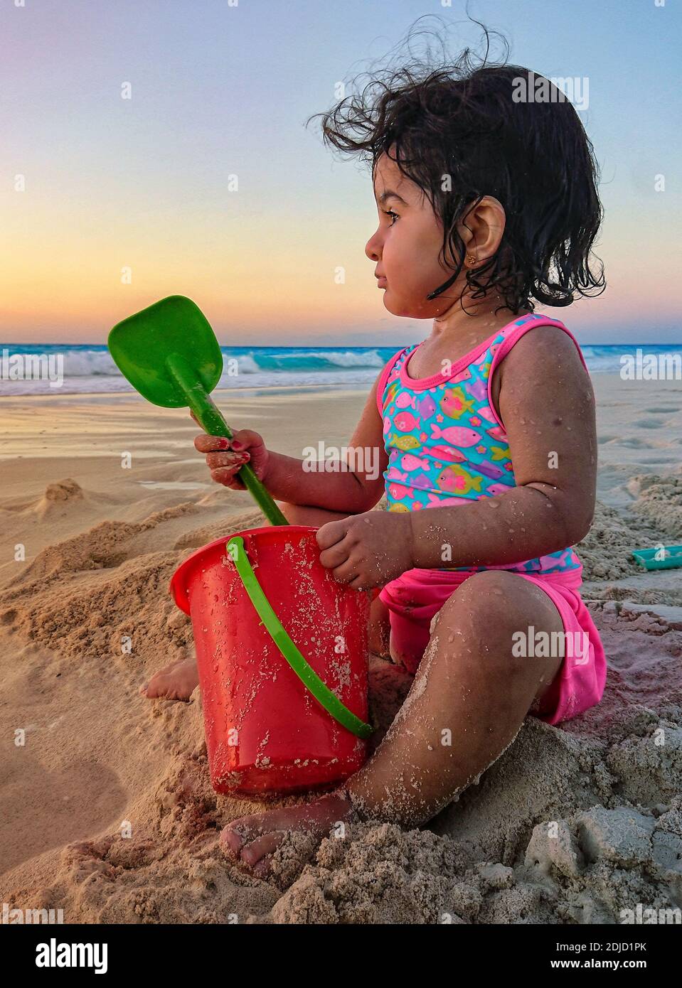 Petite fille mignonne qui regarde loin en s'asseyant à la plage contre le  ciel pendant le coucher du soleil Photo Stock - Alamy