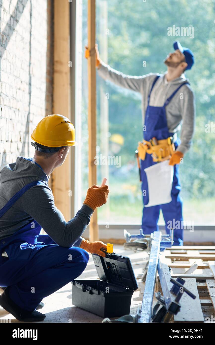 Un jeune ingénieur de sexe masculin portant une combinaison et un casque de sécurité travaillant sur le chantier de construction de chalets, cueillant l'instrument de la boîte à outils avec un collègue flou en arrière-plan, par une journée ensoleillée. Concept de bâtiment Banque D'Images