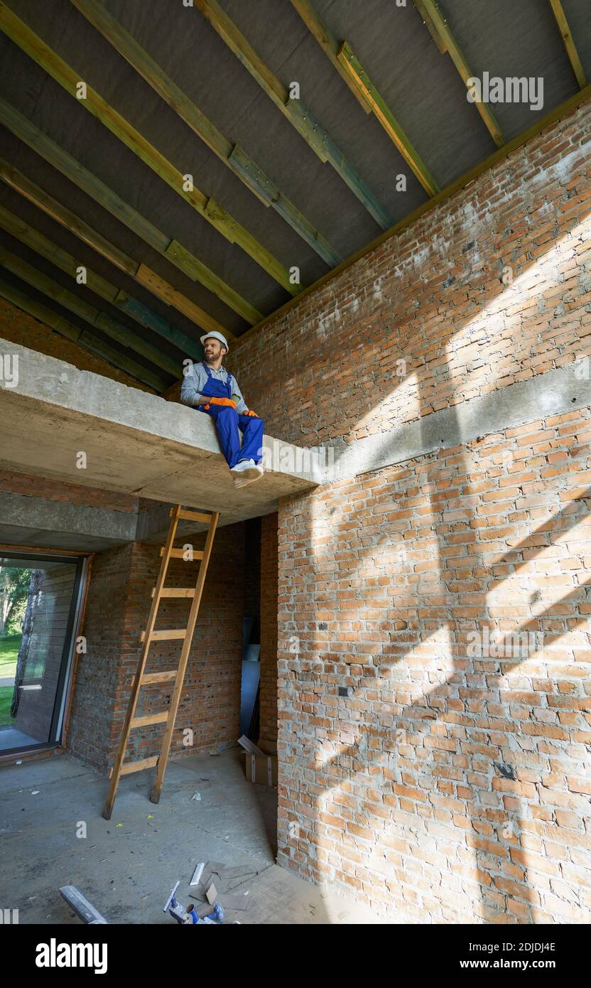 Joyeux jeune homme constructeur dans des combinaisons bleues et un casque dur assis sur le sol en béton, prenant une pause tout en travaillant sur la construction de la maison. Maison de bâtiment, concept de profession. Vue à angle bas Banque D'Images