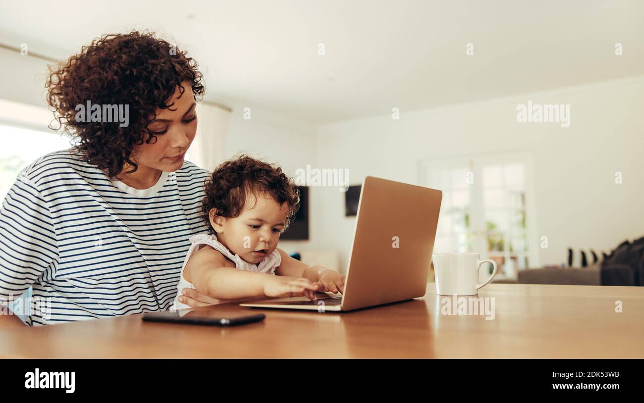 Mère regardant bébé appuyant avec les touches du clavier de l'ordinateur portable. Femme indépendante assise avec bébé et ordinateur portable à la maison. Banque D'Images