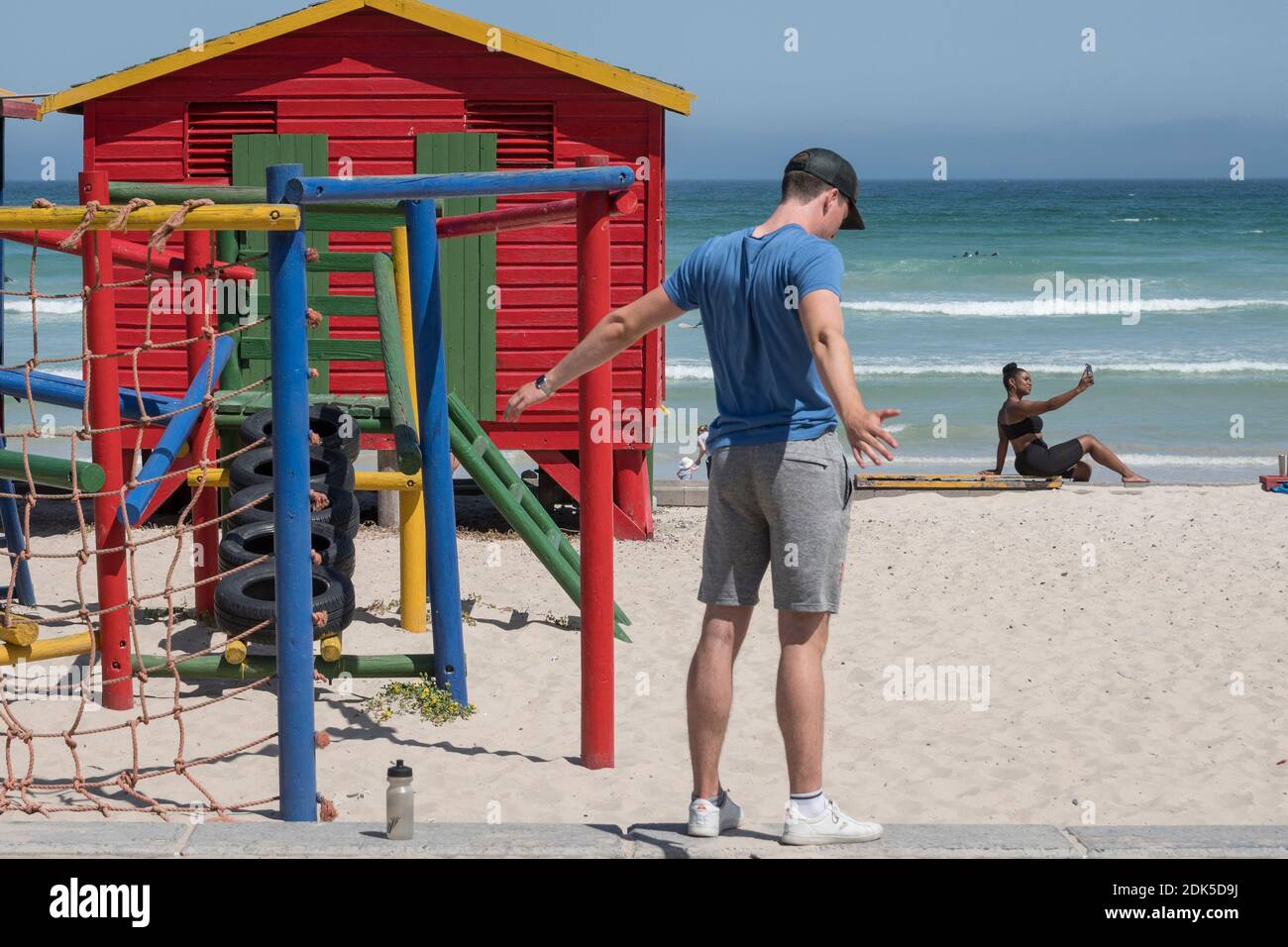 Un jeune homme s'exerce et une femme prend un selfie de téléphone portable à Surferss' Corner, Muizenberg Beach, False Bay, Cape Town, Afrique du Sud. Banque D'Images