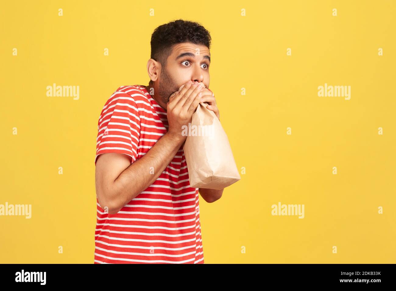 Portrait de profil homme effrayé en t-shirt rayé rouge exhalant et inhalant dans l'emballage, en utilisant le sac en papier pour améliorer le bien-être, surmontant la peur de stress. Banque D'Images