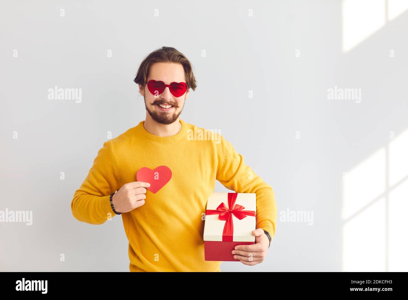 Homme joyeux dans des verres festifs tenant un cadeau et un coeur comme un symbole de l'amour pour son bien-aimé. Banque D'Images