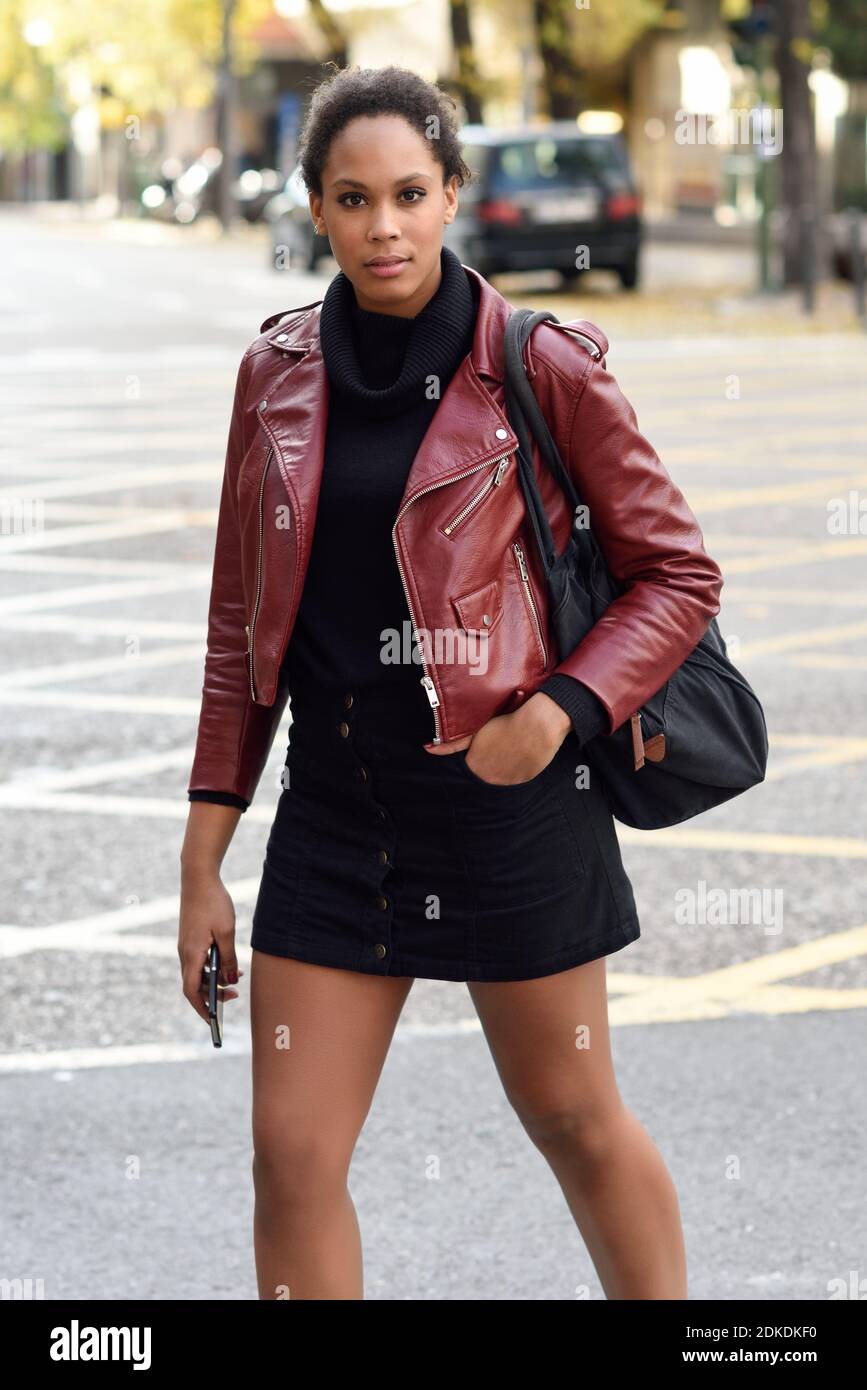 Jeune femme noire avec afro hairstyle debout dans une rue urbaine. Femme  portant mixte veste en cuir rouge, noir pull et jupe avec arrière-plan de  la ville. Sac Femme et smartphone Photo