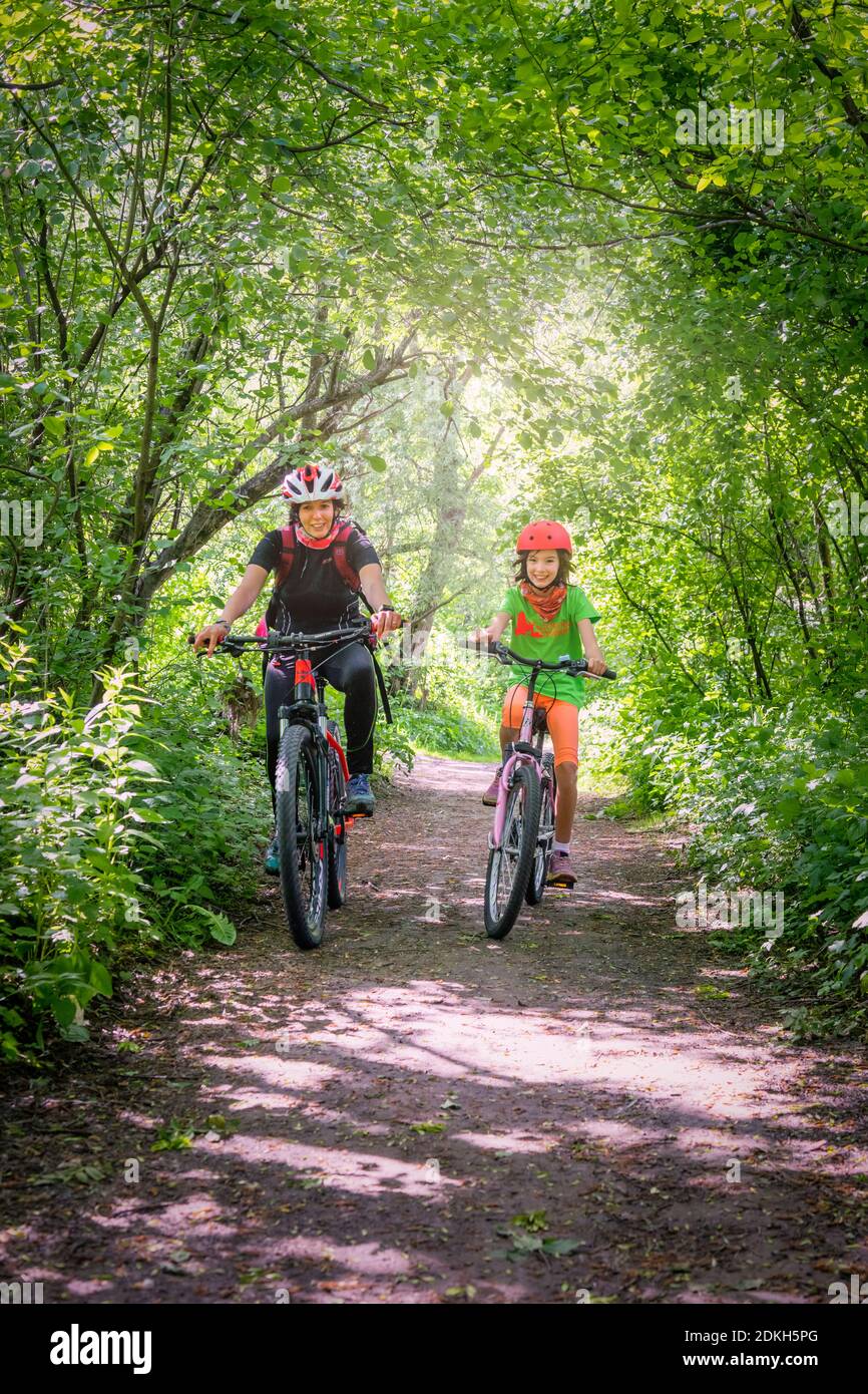 L'Italie, la Vénétie, Belluno, Agordino, maman souriante et fille à vélo sur une route parmi les arbres Banque D'Images