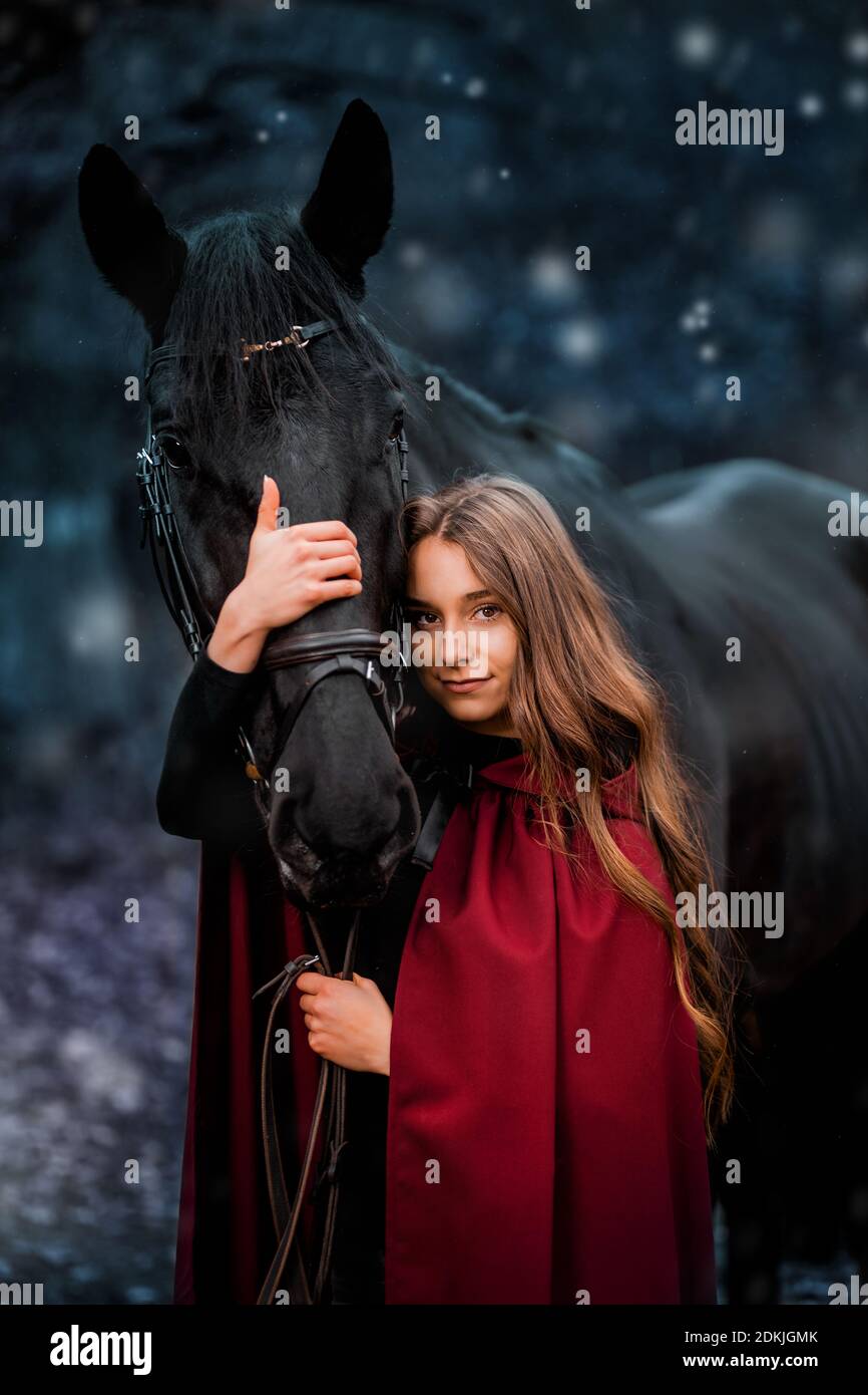 Portrait fabuleux d'une belle jeune femme aux longs cheveux bruns, suite au manteau rouge ou bordeaux avec son cheval noir en hiver Banque D'Images