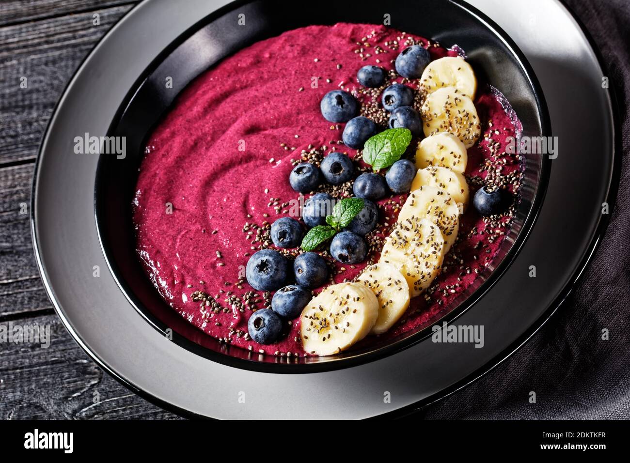 Bol à smoothie aux myrtilles et aux bananes avec yaourt, noix de cajou, graines de chia et graines de lin sur une assiette noire sur fond de bois rustique foncé avec ingrédient Banque D'Images