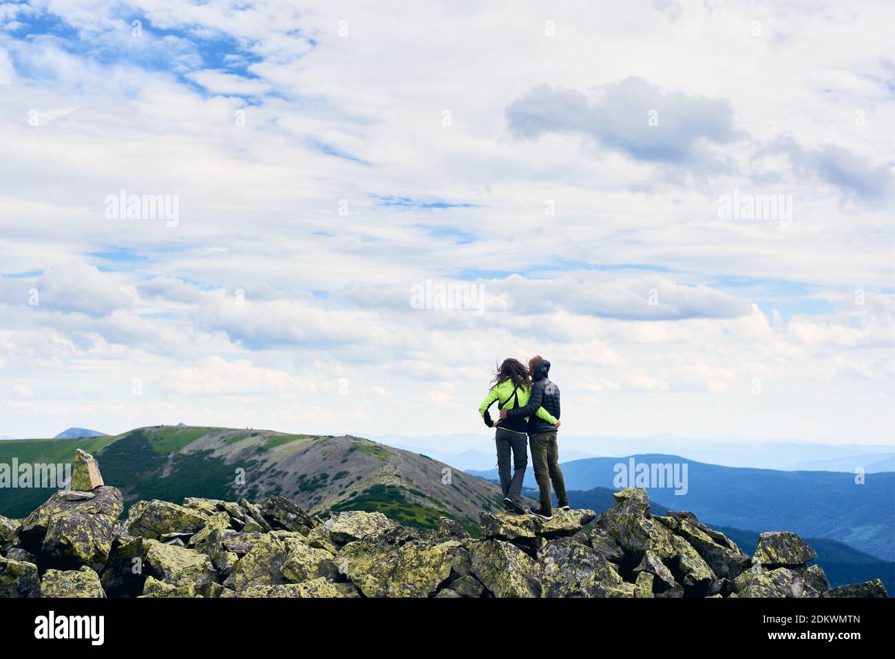 Vue arrière les jeunes sont debout dans embrasser sur des pierres au sommet avec belle vue vallée Carpathian montagnes. Fille et gars portant des vestes sous ciel nuageux par temps venteux. Mode de vie actif Banque D'Images