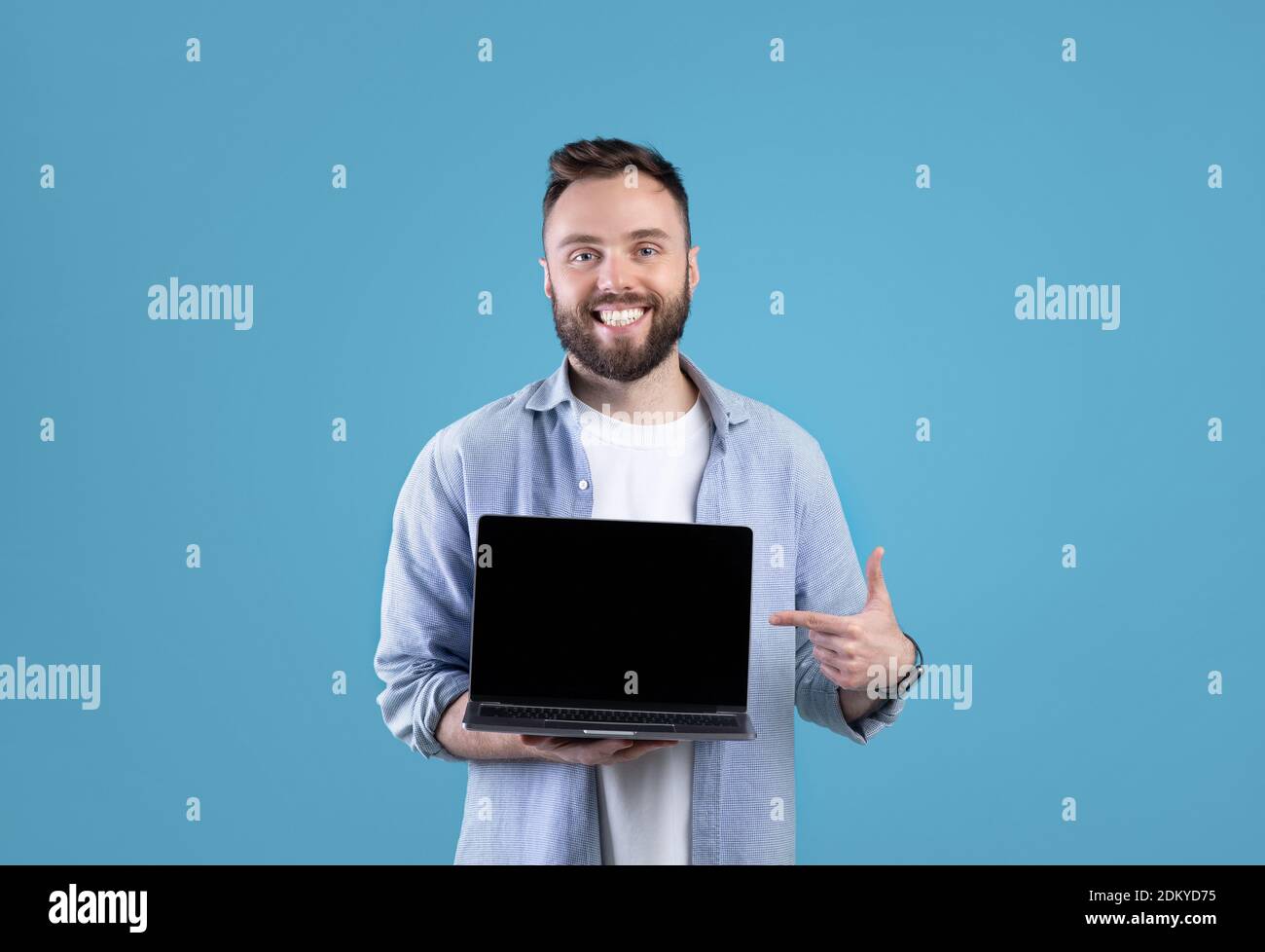 Portrait d'un homme à barbe heureux tenant un ordinateur portable avec écran vide sur fond bleu studio, maquette pour le design Banque D'Images