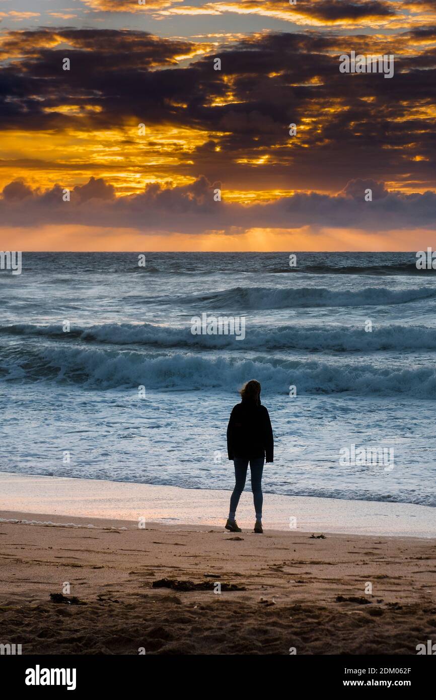 Une femme debout seule sur la plage de Fistral silhouetée par un coucher de soleil spectaculaire à Newquay en Cornouailles. Banque D'Images