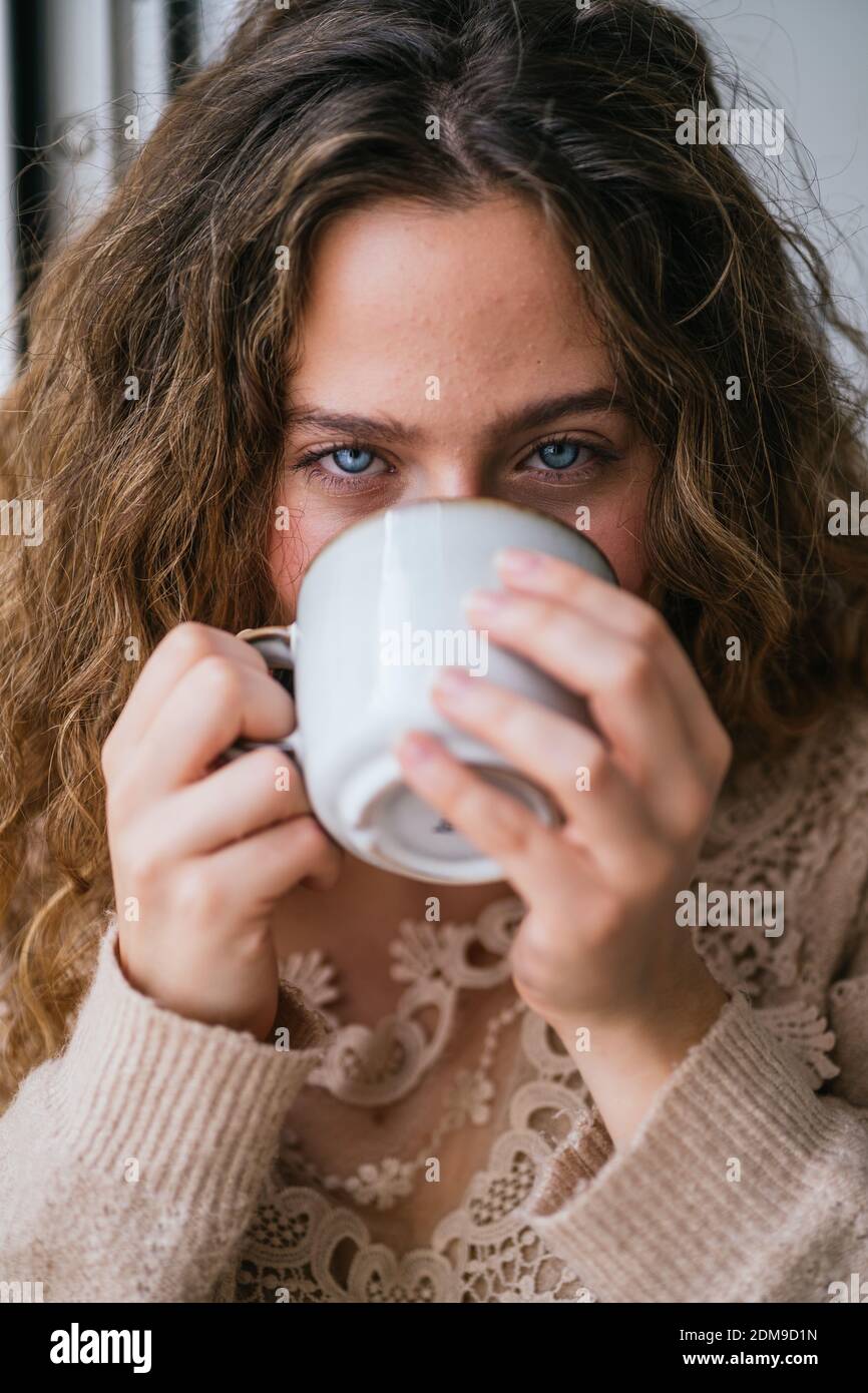 Jeune femme aux yeux bleus sirotant une boisson chaude et regardant à l'appareil photo Banque D'Images