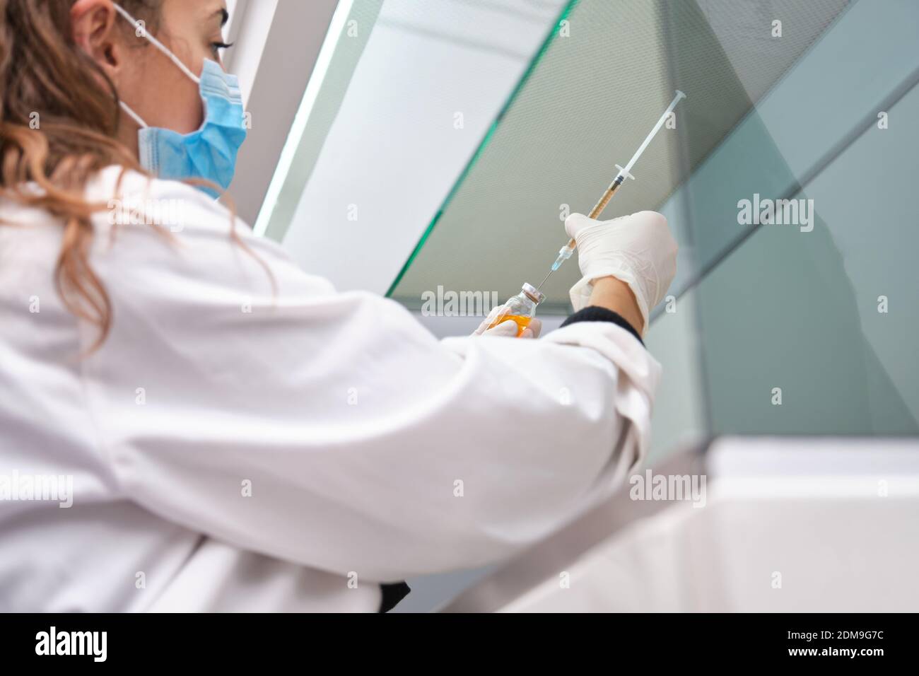 Une jeune scientifique féminine portant un masque facial tenant un flacon, une seringue et une aiguille de vaccin coronavirus dans une hotte à flux laminaire. Développement du vaccin Covid-19. Banque D'Images