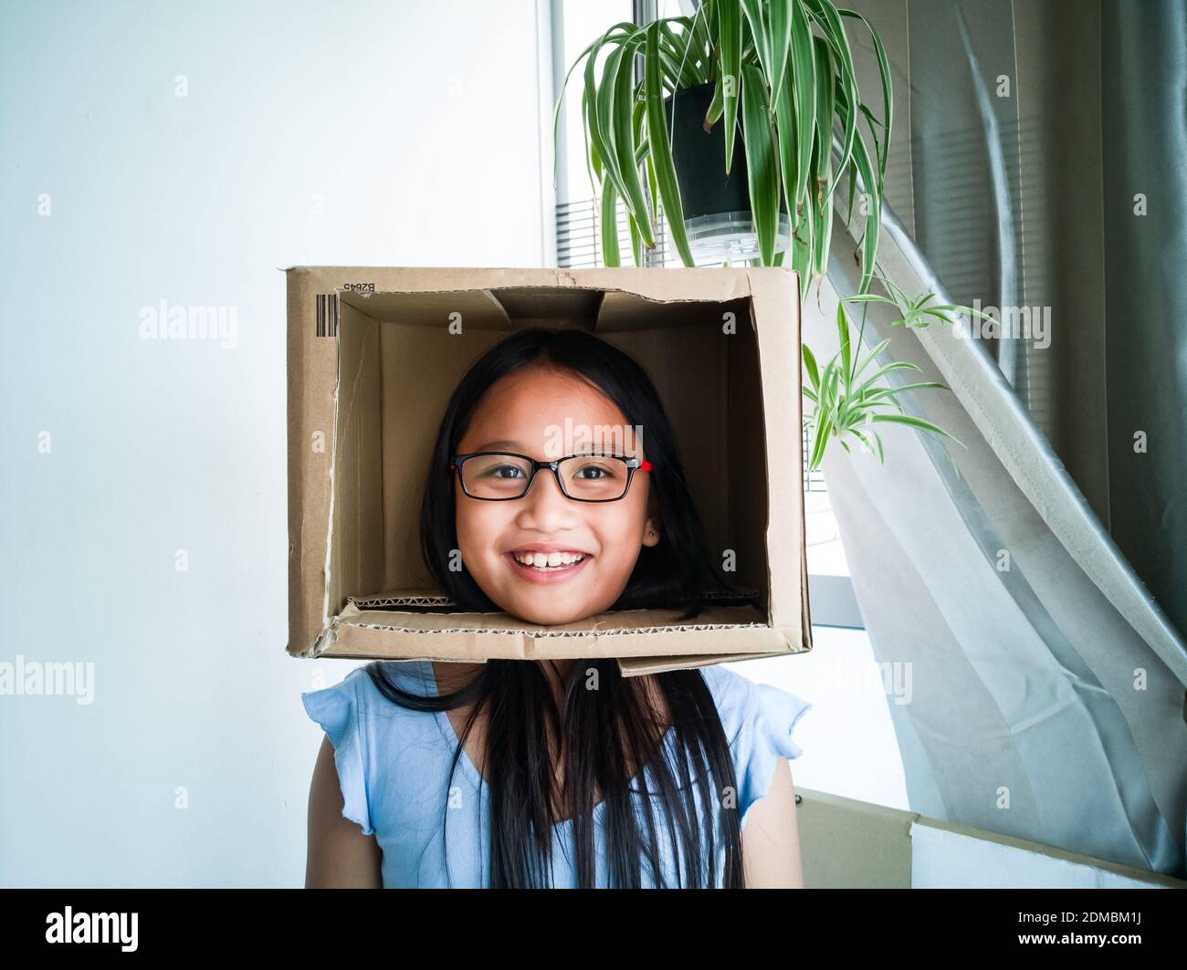 Drôle asiatique jeune fille portant des lunettes de soleil portant une boîte  de carton sur sa tête. Faire semblant de jouer comme un nouveau-scaster à  la maison pendant le verrouillage du covid