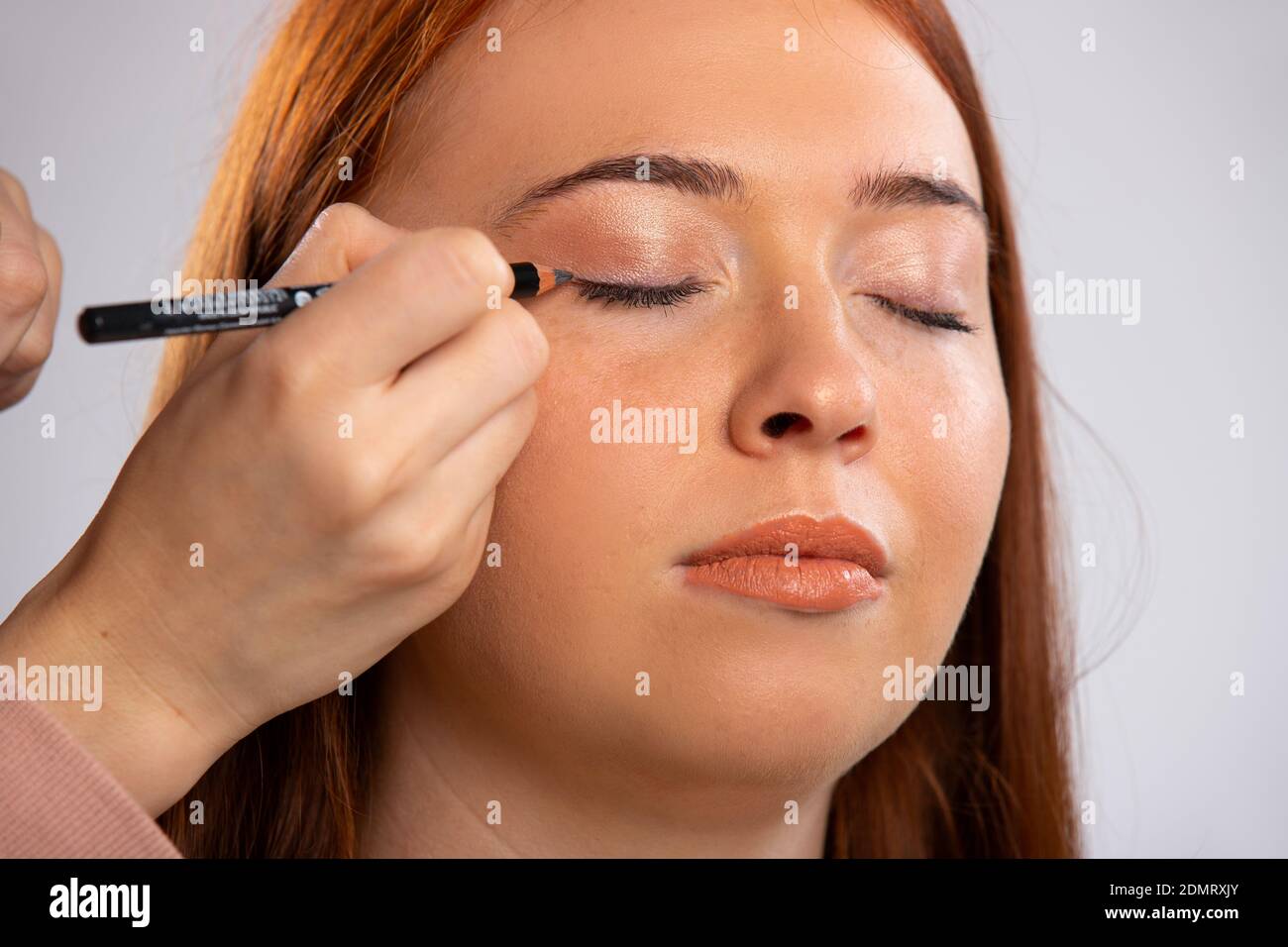Gros plan photo de maquillage pour une fille aux cheveux rouges, yeux fermés et un crayon cosmétique pour les yeux. Concept de beauté. Banque D'Images