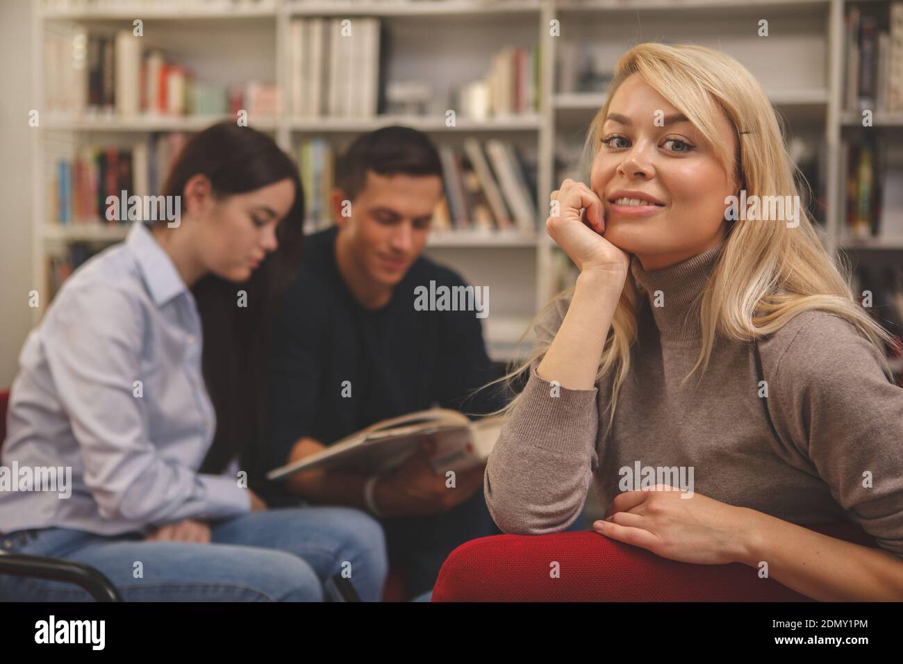 Gorgeous happy blond haired woman smiling to the camera, relaxing at college library with her friends tudying on the background. Lovely female student Banque D'Images