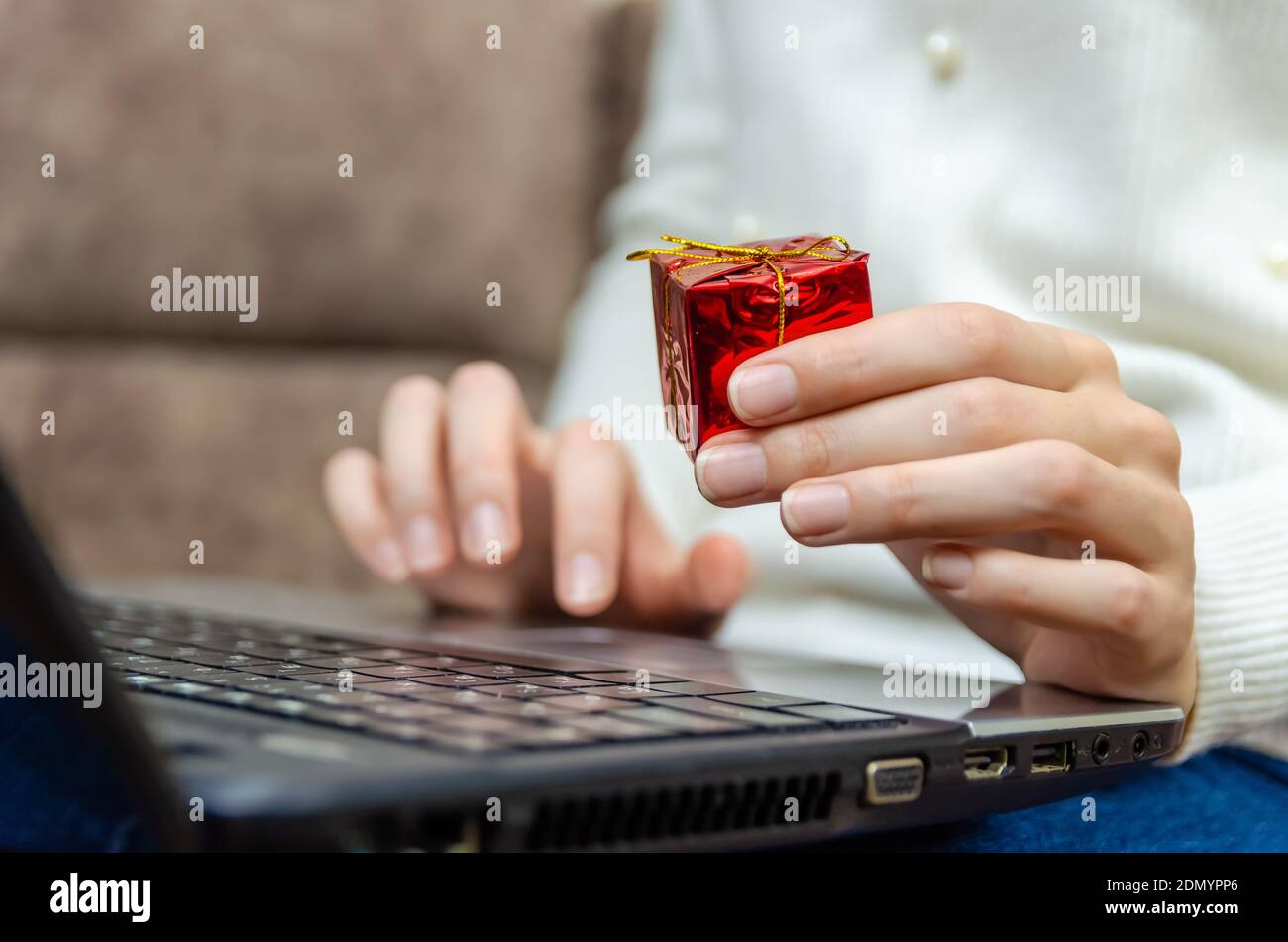 Une femme dans un chandail blanc tient un ordinateur portable sur ses genoux et tient une boîte cadeau rouge dans sa main en gros plan. Concept de Noël, nouvel an, vacances Banque D'Images