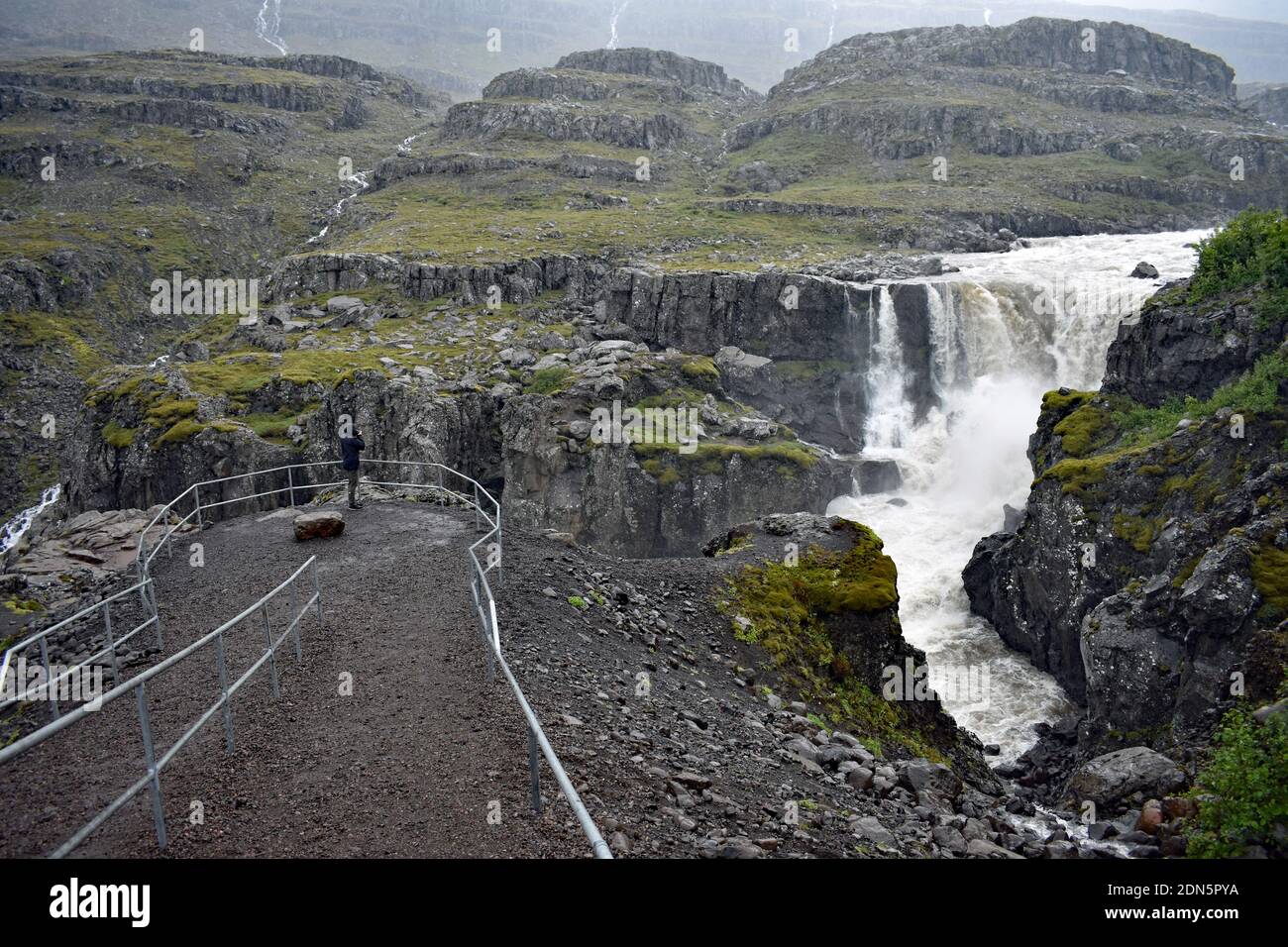 Un homme portant des mains noires se tient derrière des rampes dans la zone d'observation de Nykurhylsfoss (Sveinsstekksfoss) près de Djupivogur dans les Eastfjords d'Islande. Banque D'Images