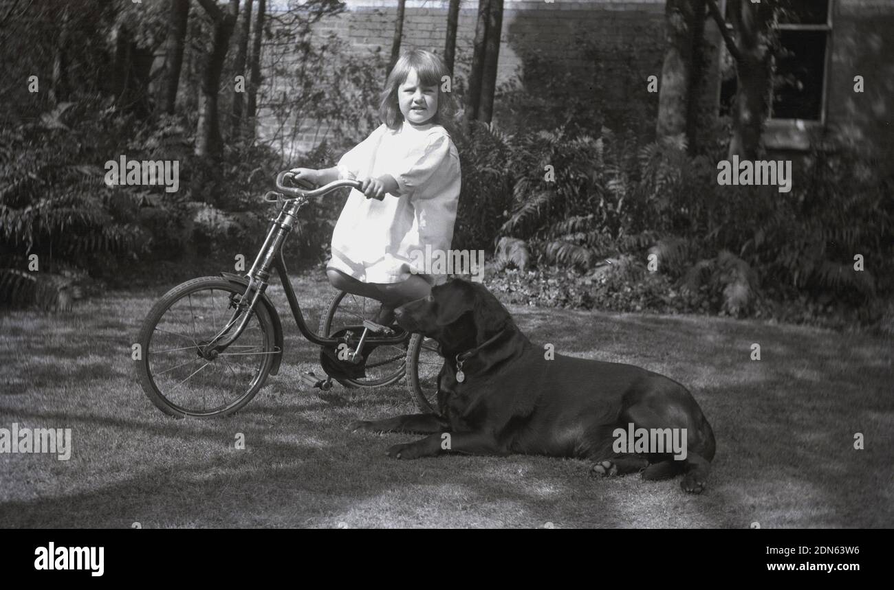 1920s, historique, à l'extérieur d'un jardin où la loi est passée, une jeune fille portant une grenouille d'été en coton assise sur son tricycle à cadre en acier, avec son chien de compagnie couché à côté d'elle, Angleterre, Royaume-Uni. Banque D'Images