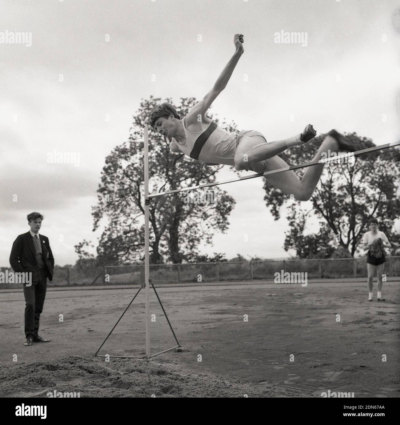 1965, à l'extérieur, dans un domaine sportif, sous la surveillance d'un élève, un jeune homme faisant le saut en hauteur lors d'une journée de sport au lycée, en utilisant une forme de la méthode de la selle, la selle parallèle, lorsque la jambe principale est en haut dans l'air et que la tête et le tronc se détalèrent de la barre en même temps. Le champion olympique de 1956, Charles Dumas - et le premier cavalier à effacer 7ft - a utilisé cette version de la selle. L'autre version de la selle est la méthode de plongée, où la tête passe au-dessus de la barre en premier. Dans amateur saut élevé les deux styles sont souvent mélangés ensemble, dans un ce qui vous fait jamais au-dessus des travaux de barre. Banque D'Images