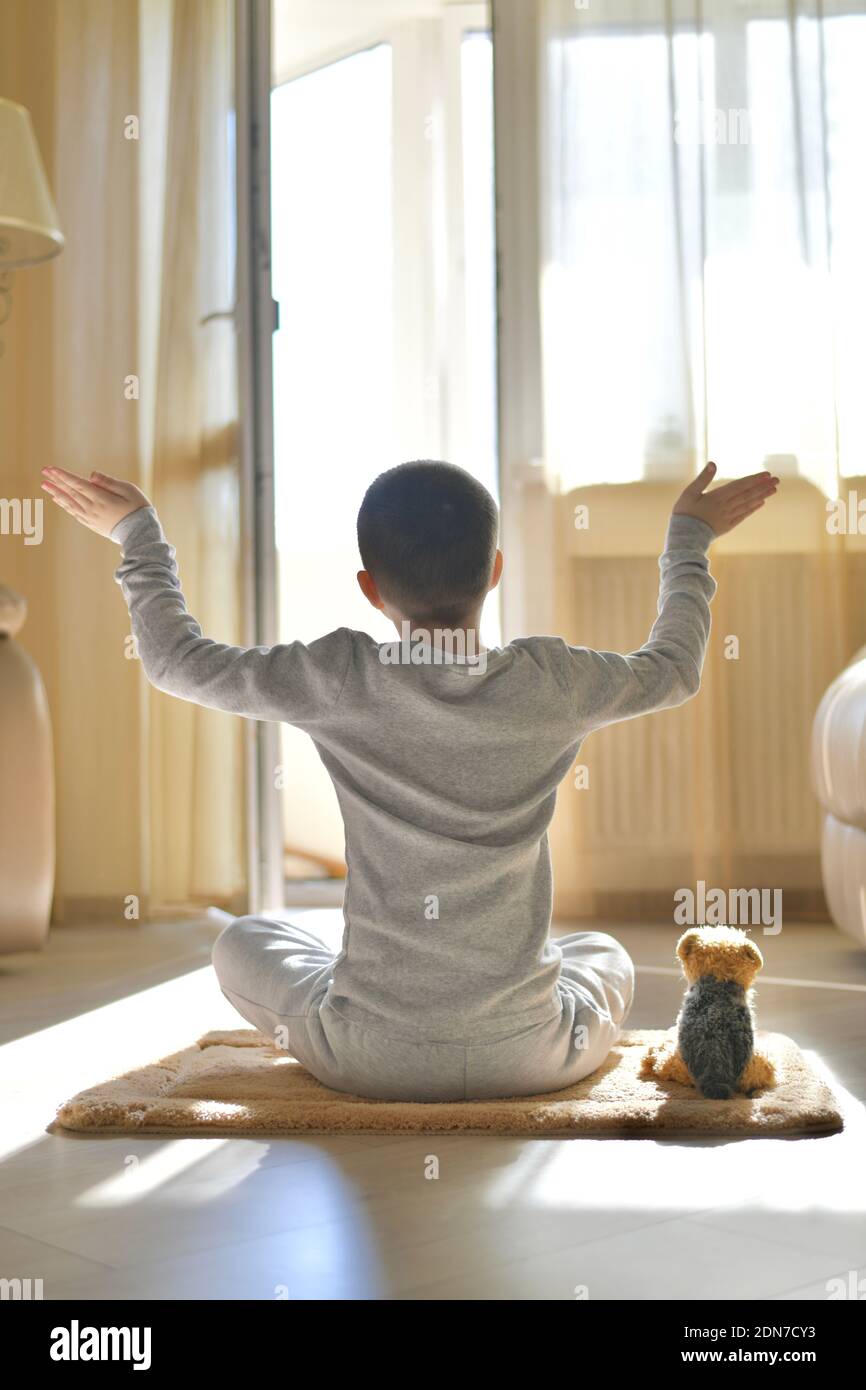 L'enfant s'assoit avec son dos à la maison. Sport de fitness yoga sur le tapis avec peluche chien. Méditation dans la position du lotus avec les bras ouverts, sur le sol Banque D'Images