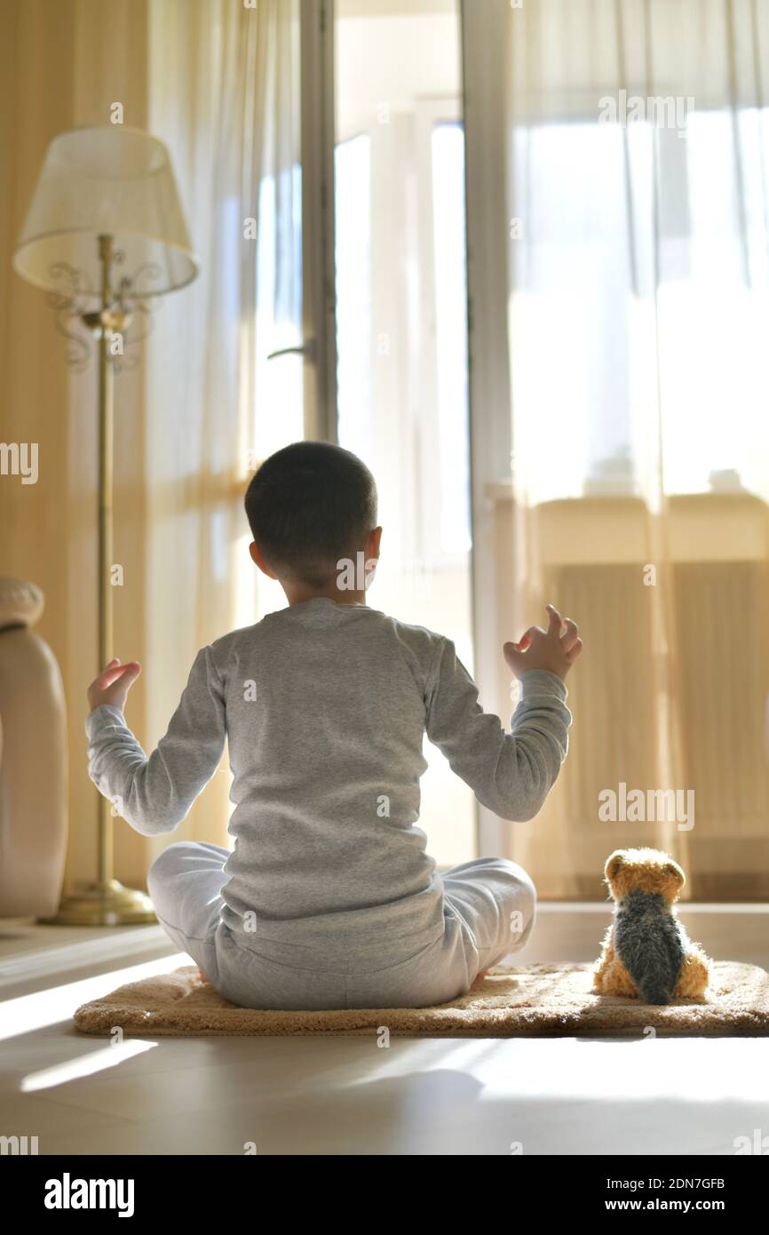 L'enfant s'assoit avec son dos à la maison. Entraînement physique yoga sur le tapis avec peluche chien. La méditation dans la position du lotus avec les mains fait mudra, sur le floo Banque D'Images