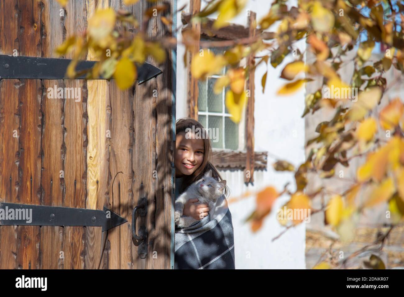 Une fille souriante avec un chat blanc regarde de derrière une porte de grange rustique en bois dans un jardin d'automne Banque D'Images